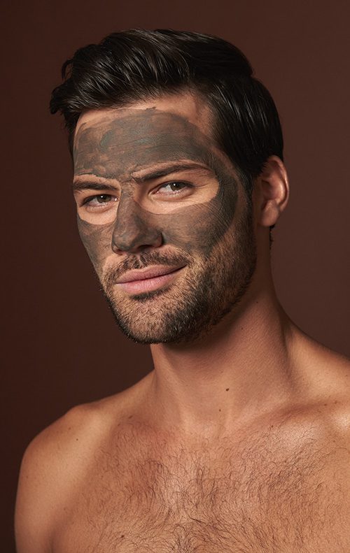 Beauty procedure and skin care. Close up portrait of young self-confident smiling man with black face mask looking straight on camera. Isolated on brown