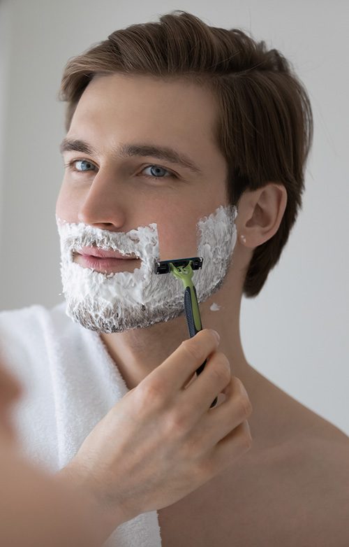 Positive attractive young guy with gel foam on lower face shaving with razor at mirror. Handsome man enjoying morning bath routine, caring for appearance, healthy facial skin. Hygiene, skincare concept