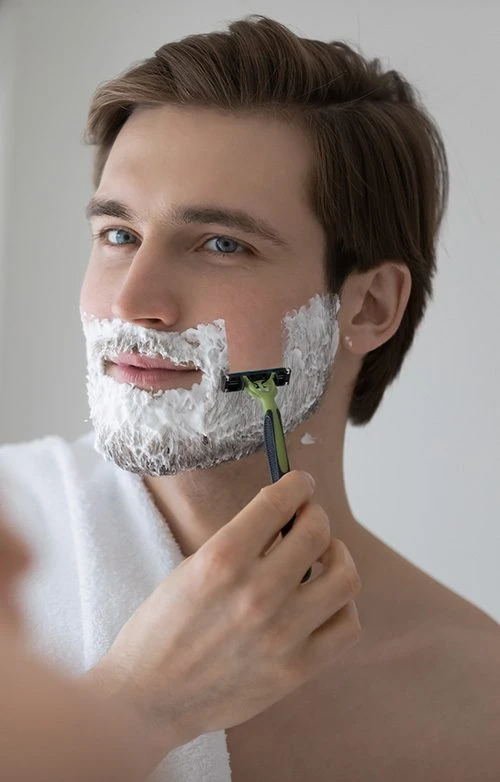 Positive attractive young guy with gel foam on lower face shaving with razor at mirror. Handsome man enjoying morning bath routine, caring for appearance, healthy facial skin. Hygiene, skincare concept