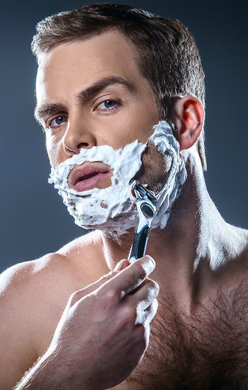 Studio portrait of handsome young man. Man with naked torso and shaving foam on face looking at camera and shaving