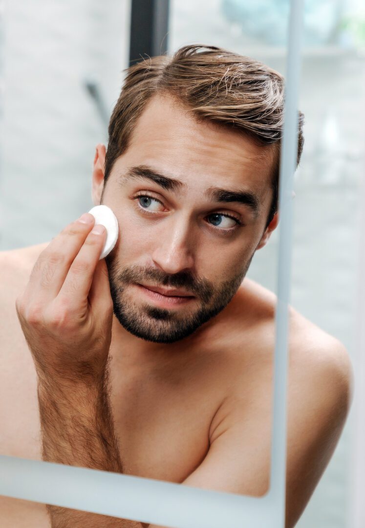 Handsome young shirtless man applying aftershave lotion with a cotton pad while standing at the bathroom mirror